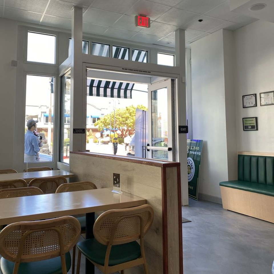 Interior of Jack's Bao, showing light wood tables, wicker chairs, a green booth, and large windows.