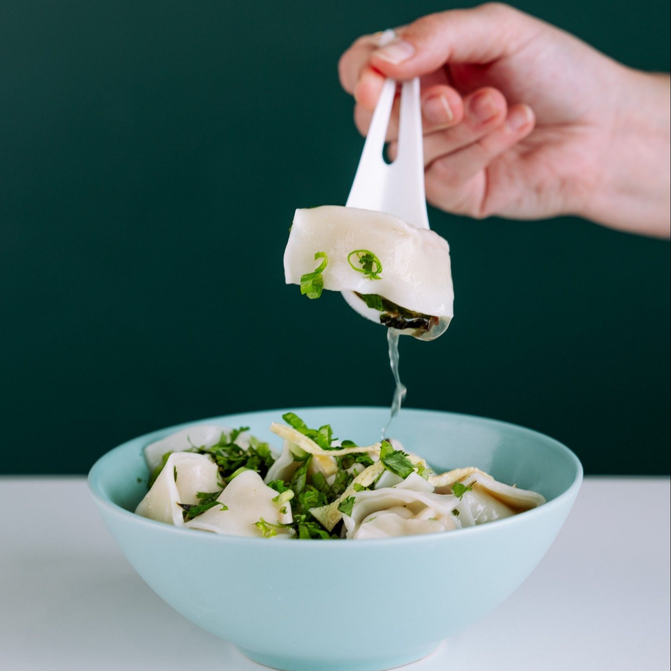 A hand lifts a dumpling with a white spoon from a bowl of dumplings and broth.